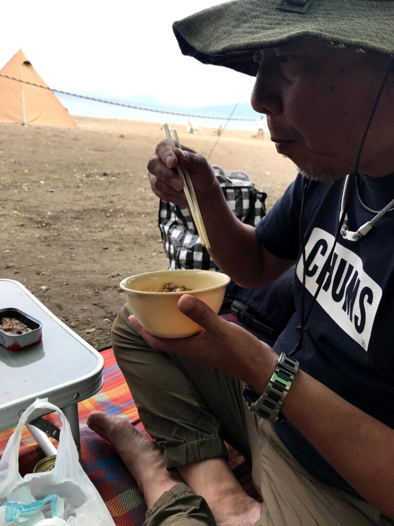 ハイエースのそばでキャンプ飯を食べる男性のアップ(支笏湖畔)