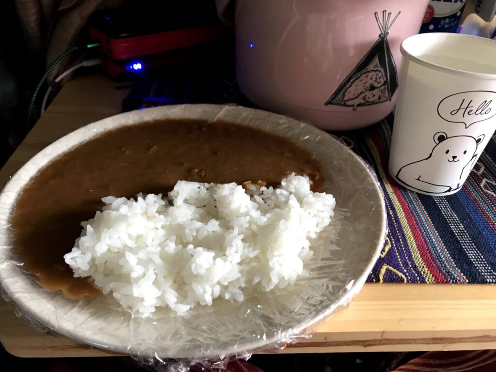 車中泊の車内で準備されたレトルトカレーと白ごはんの皿、紙コップや炊飯器も写る