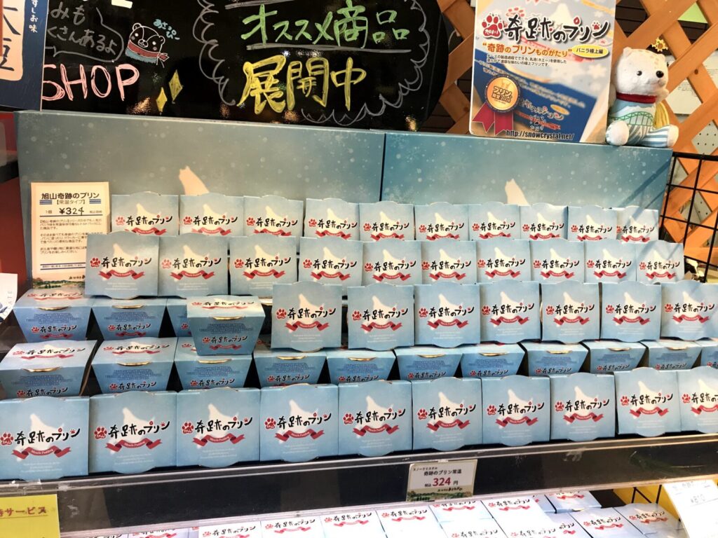 旭川の道の駅で販売されている「奇跡のプリン」の陳列棚。水色のパッケージがずらりと並び、上部にはポップとぬいぐるみが置かれている。