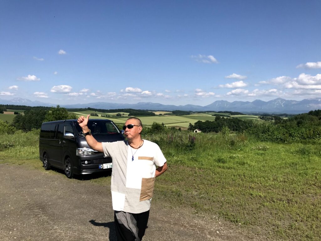 北海道美瑛の丘でハイエースの前に立ち、親指を立てる男性のポーズ