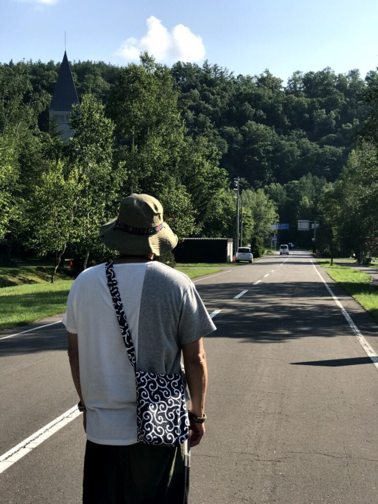 帽子をかぶった男性が振り返るように立ち、奥に塔と緑の山々が広がる。