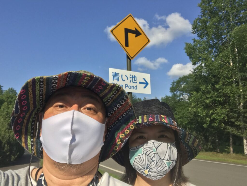 「青い池」の案内標識前で撮影した、帽子とマスク姿の男女のツーショット