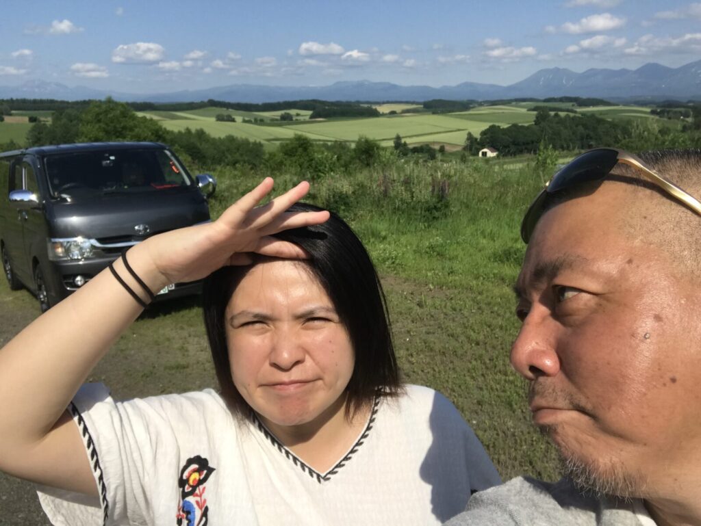 女性がまぶしそうに手をかざし、男性が見つめる美瑛の丘での自撮り写真。