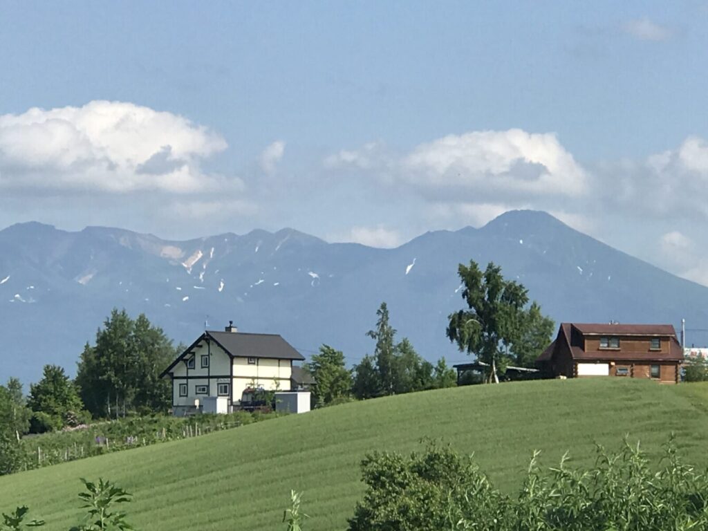 美瑛の丘に立つ洋風の家と、背景に連なる十勝岳の山々。