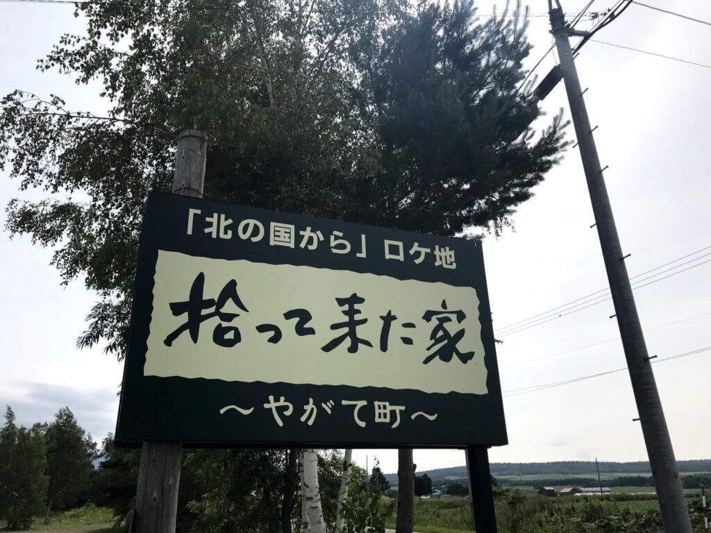 「北の国から」ロケ地「拾ってきた家 ～やがて町～」の大きな看板が、空と田園風景を背景に立っている。