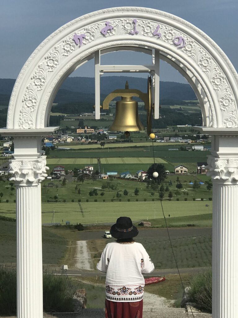 白いアーチと鐘の前に立つ女性の後ろ姿。遠くには田園風景が広がる