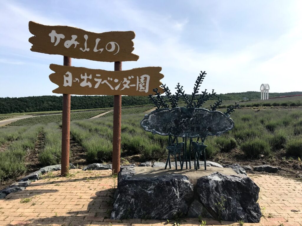 「かみふらの 日の出ラベンダー園」と書かれた木製の看板と、ラベンダー畑が広がる丘の風景