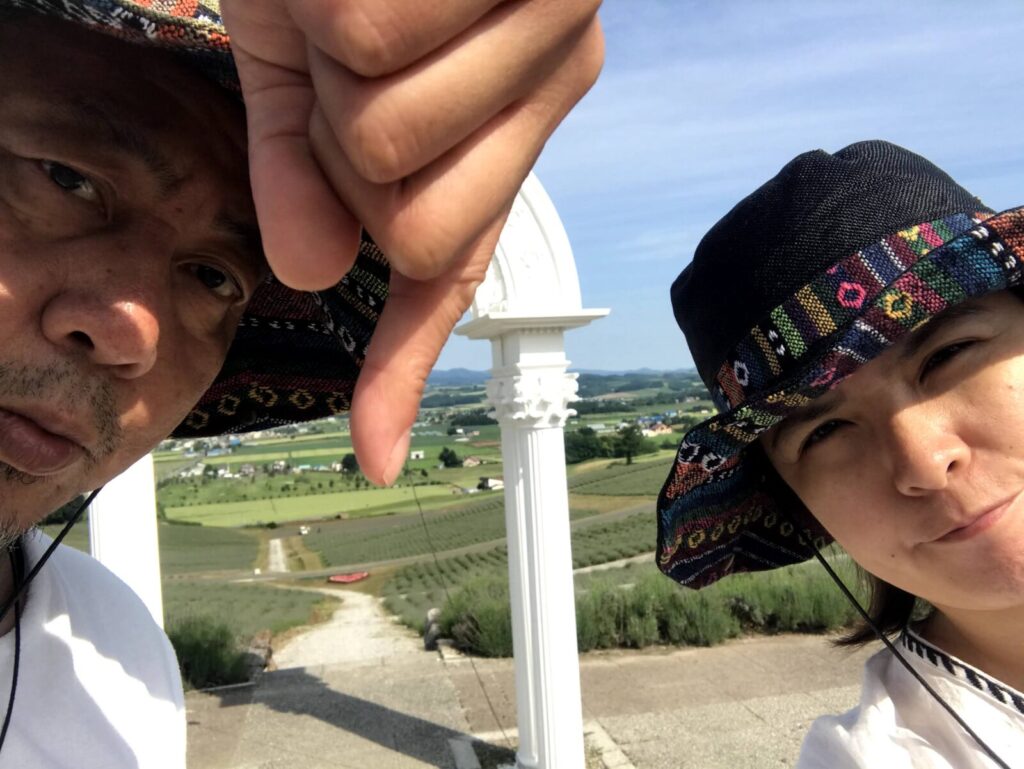 ラベンダー畑を背景に、親指を下に向けて不満げな表情を浮かべる帽子姿の男女の自撮り写真