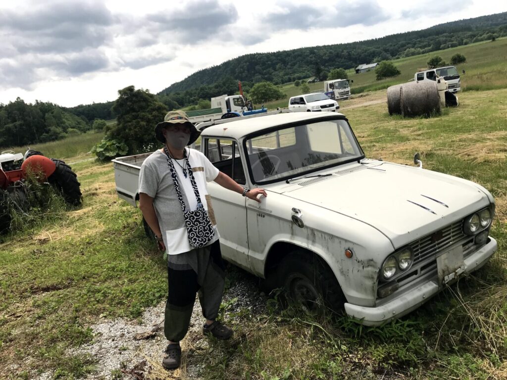 北の国からのロケ地で撮影された、旧車と男性の記念写真。背景には牧草地や作業車が並ぶ。