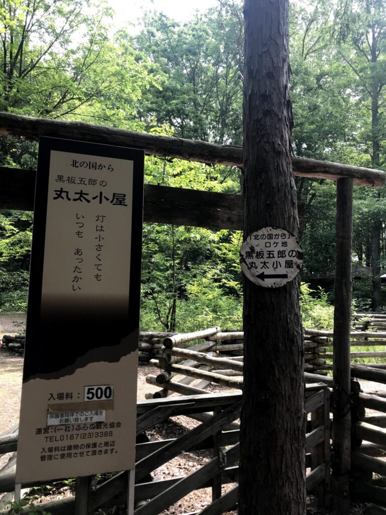 「北の国から」黒板五郎の丸太小屋への案内板。木の柵や看板が森の中に立っている。