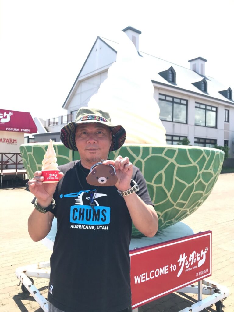 ポプラファームの巨大メロンソフトオブジェの前でソフトクリームとクマポーチを持つ男性。