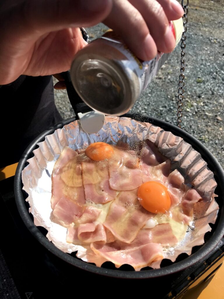 車中泊の朝食でベーコンと卵に調味料をかけて調理中