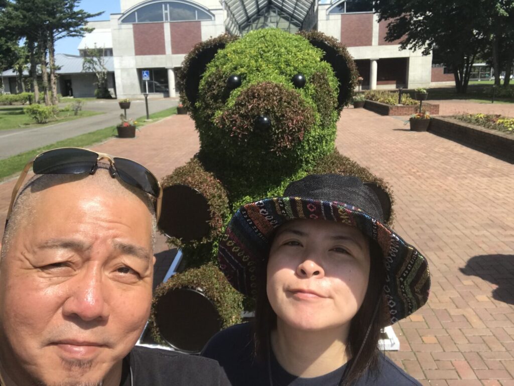 道の駅しかおいのクマオブジェの前で記念撮影をする夫婦。背景に施設が見える。