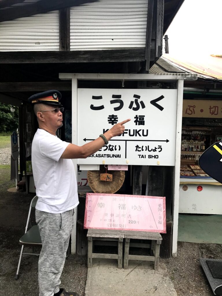幸福駅の駅名標の前で駅長帽をかぶってポーズを決める男性