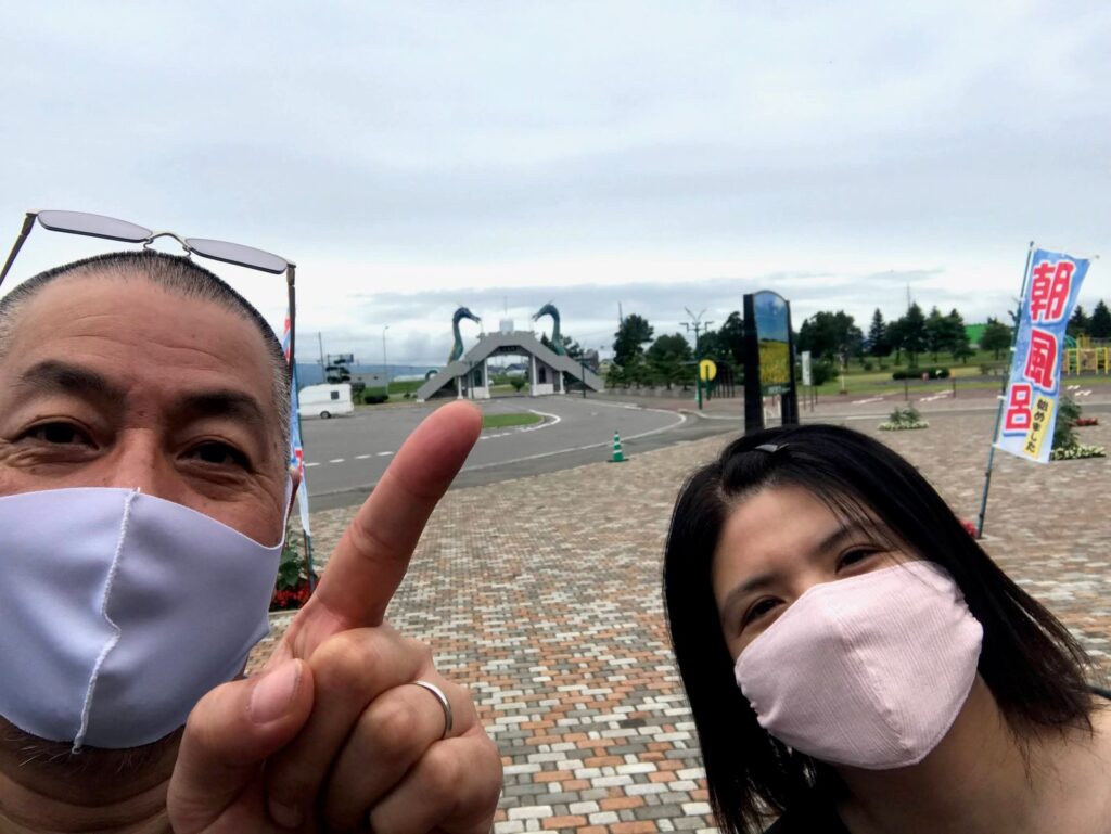 「朝風呂」ののぼりの前で指をさす旦那とくま嫁の自撮り。奥に竜の門